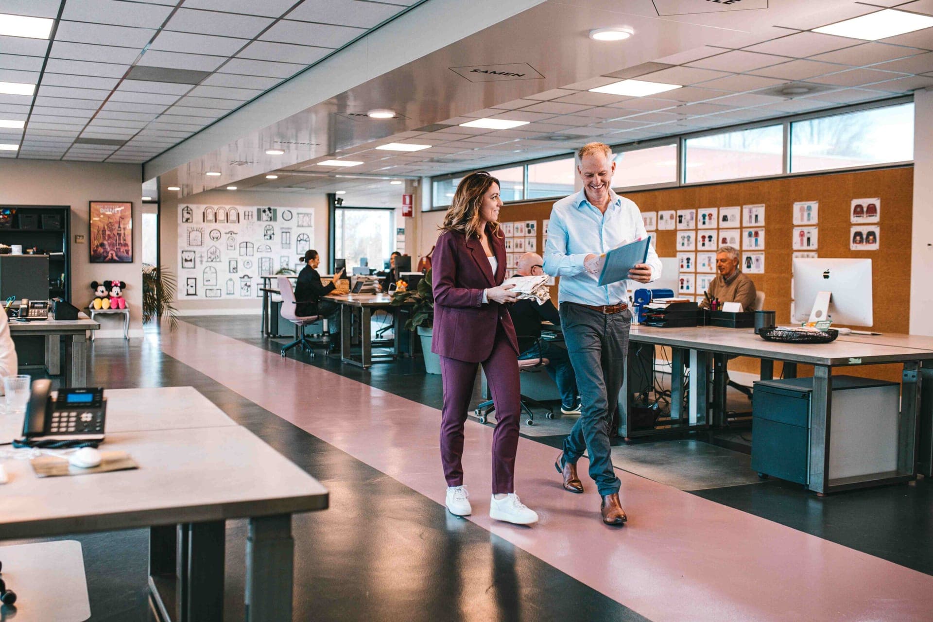 Two colleagues walking through the open-plan design office with a folder and clothing samples, garment sketches on the wall behind them