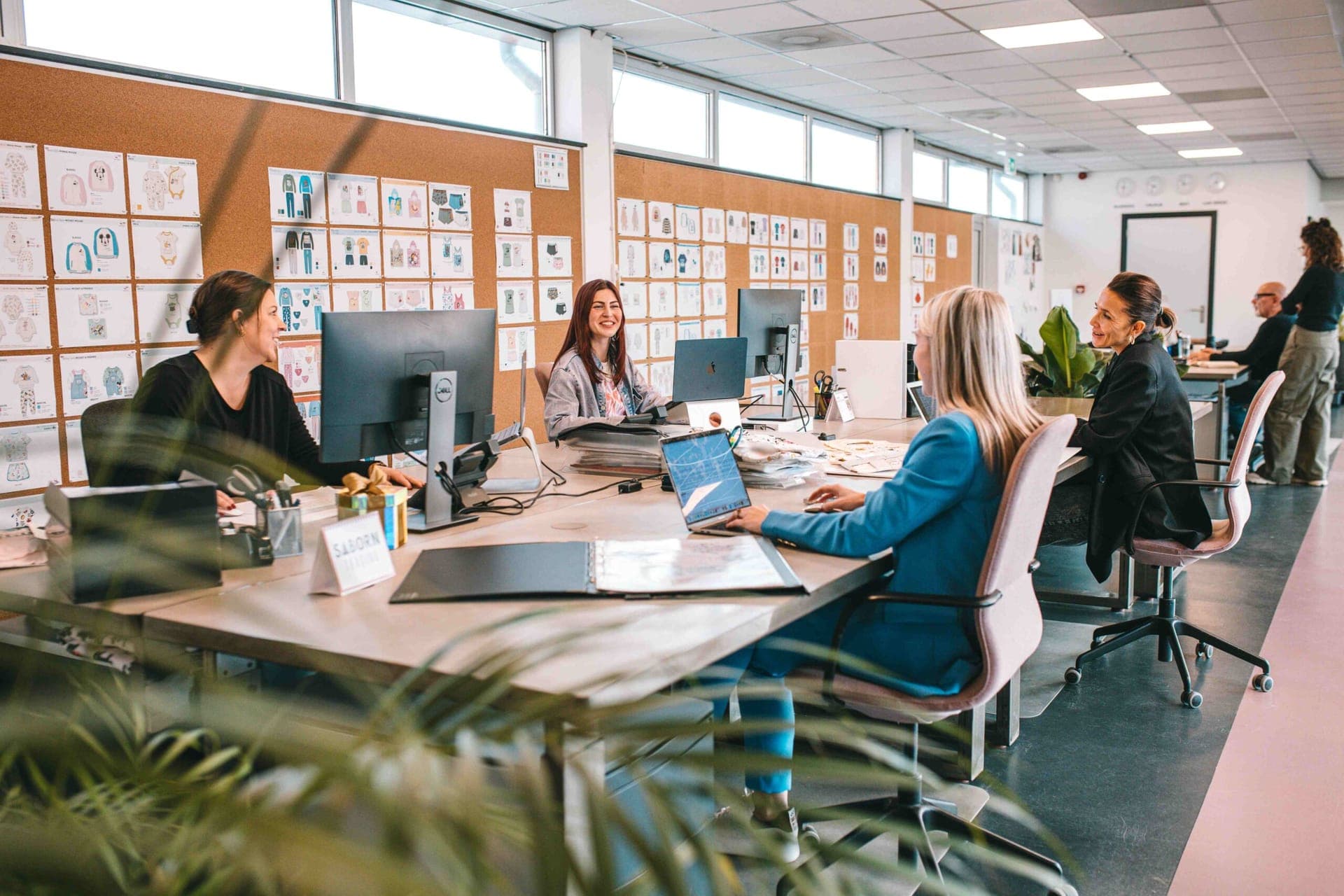 Das Designteam arbeitet an einem gemeinsamen Schreibtisch vor einer Korkwand voller Entwurfsblätter für Kleidung