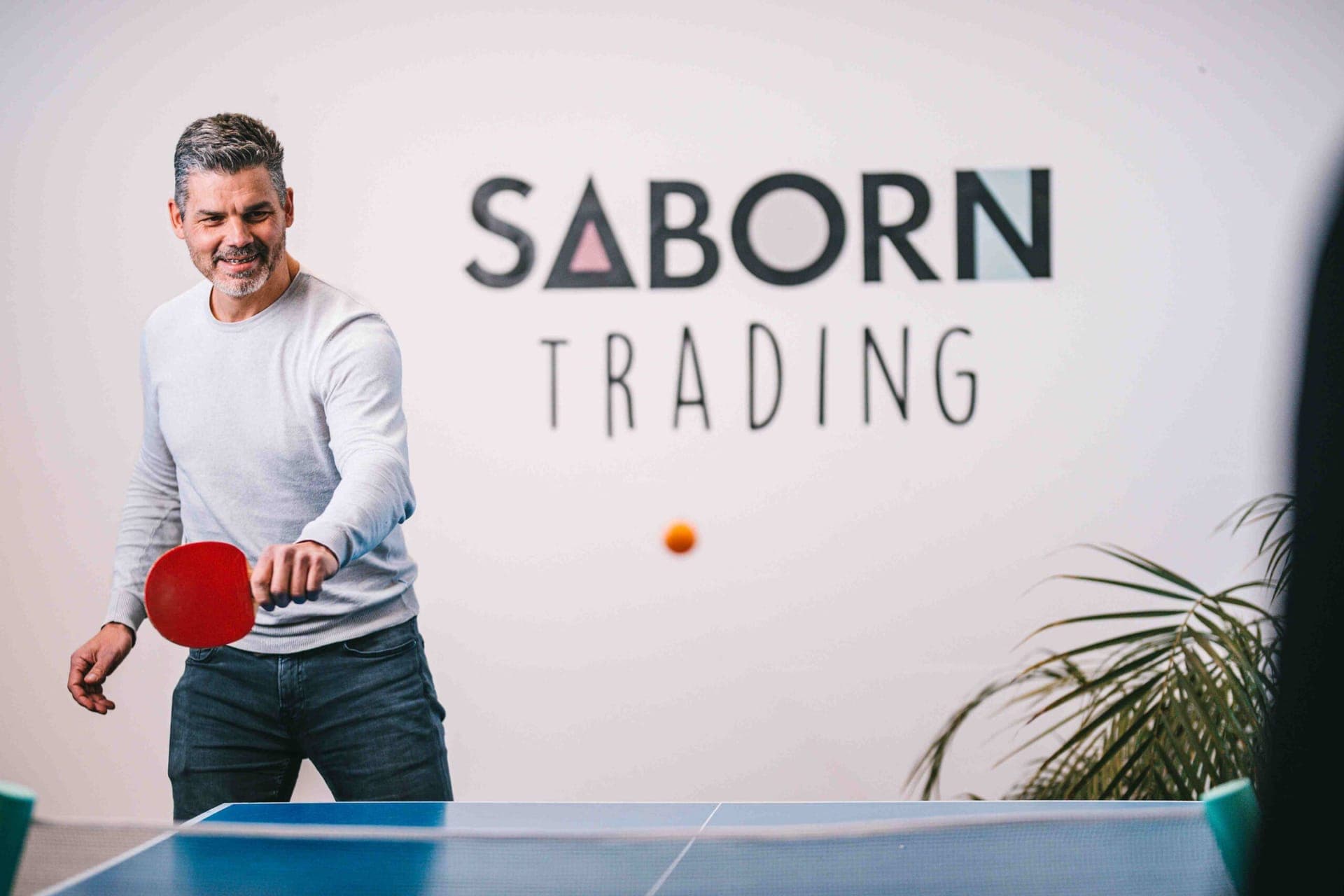Colleague playing table tennis in front of the Saborn Trading logo on the office wall