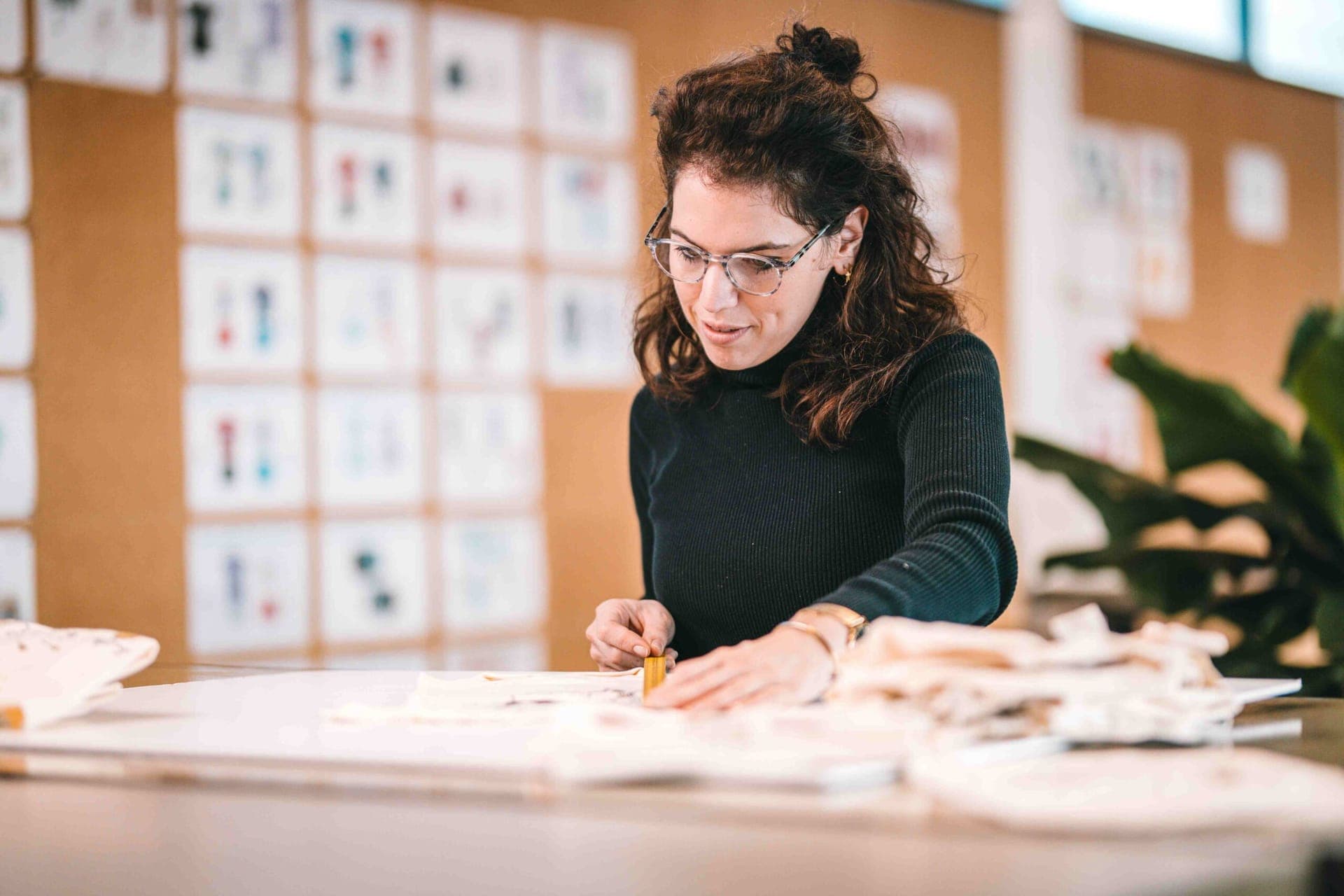 Styliste travaillant un tissu sur une table de travail, devant un mur de modèles de vêtements