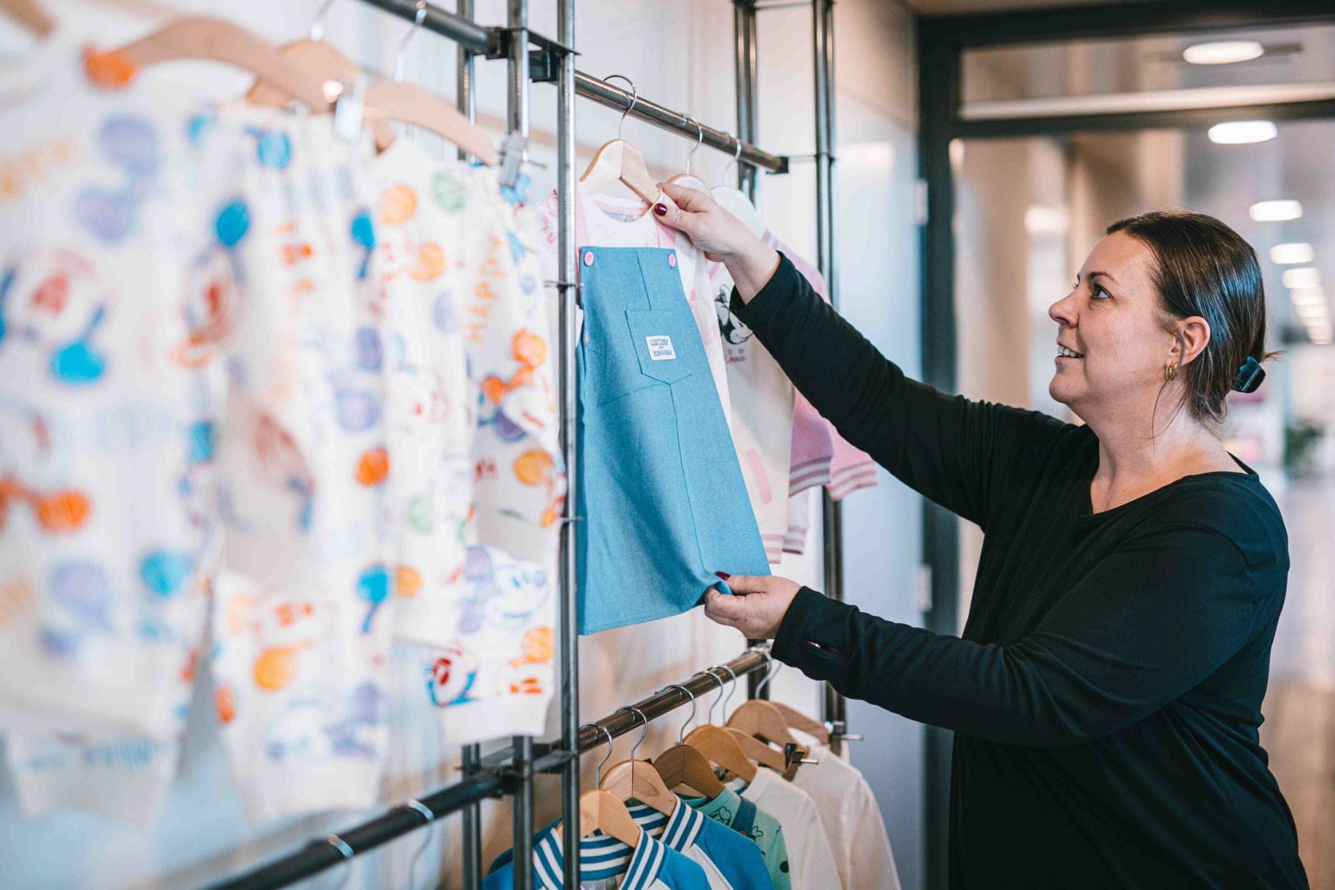 Collega hangt een denim tuinjurk aan een kledingrek naast babykleding met Mickey Mouse-print